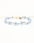 A delicate beaded bracelet featuring light blue and white flower beads with gold accents, including a small gold leaf charm, on a white background.