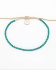 A beaded bracelet with a vibrant teal green cord and a beige adjustable sliding knot closure, featuring a small silver circular charm and wooden beads at the ends, shown on a plain white background.