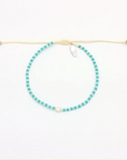 A beaded necklace with small turquoise-blue beads and a white central bead, suspended on a cream-colored adjustable cord with a small silver charm, shown on a plain white background.