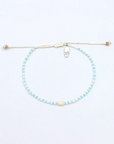 A delicate beaded cord bracelet with light blue and white beads, a central cream-colored bead, and a small silver metal charm, displayed on a clean white background.