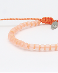 A peach-colored beaded bracelet with a matching braided cord and a small silver-toned leaf charm, photographed on a clean white background.