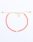 A beaded adjustable cord bracelet featuring bright orange-red round beads, a cream-colored adjustable rope closure with a knot, and a small silver-toned safety pin clasp, shown on a plain white background.