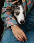 A close-up lifestyle shot of a dog peeking out from under a person's blue denim jacket. The person is wearing blue jeans and a gray patterned shirt, and the dog has a white and gray merle coat.