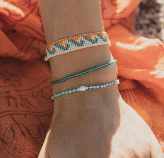 Three beaded bracelets on a person's wrist, featuring a white and orange chevron pattern, a turquoise blue beaded cord, and a turquoise and white beaded cord with a small white bead, set against an orange fabric background.