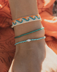 Three beaded bracelets on a person's wrist, featuring a white and orange chevron pattern, a turquoise blue beaded cord, and a turquoise and white beaded cord with a small white bead, set against an orange fabric background.