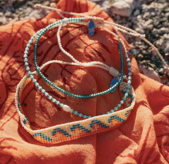 Three handmade beaded bracelets with turquoise, white, and orange details, displayed on a textured orange fabric outdoors.