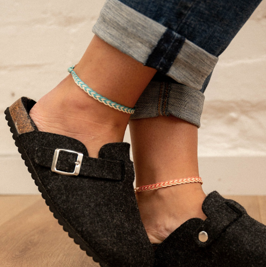 A close-up lifestyle shot of a person's feet wearing black felt slip-on clogs with silver buckles, paired with rolled-up blue jeans and two delicate braided anklets, set against a light brick wall and wooden floor.