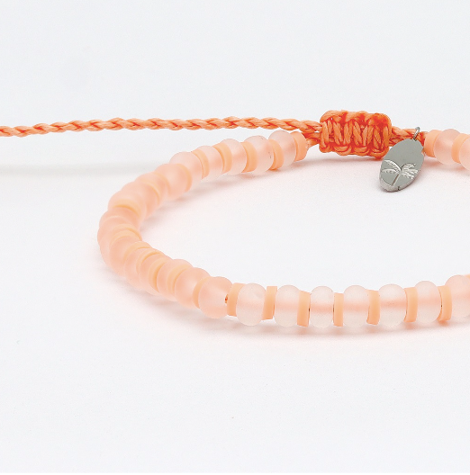 A peach-colored beaded bracelet with a matching braided cord and a small silver-toned leaf charm, photographed on a clean white background.