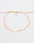 A delicate beaded bracelet with peach-pink round beads, a cream-colored adjustable cord, a small silver circular charm, and a white central bead, displayed on a clean white background.