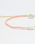 A delicate beaded bracelet with peach-pink and cream-colored beads, featuring a white shell charm, silver spacer beads, and a round silver metal charm, displayed on a clean white background.