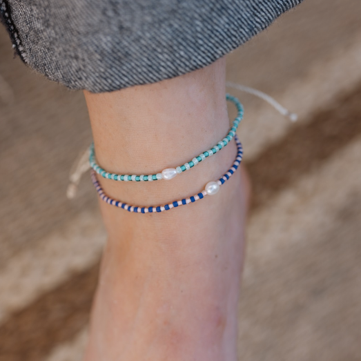 Two delicate beaded ankle bracelets on a person's ankle, one in turquoise and white, and one in navy and white, with a small white pearl accent on each. The person is wearing gray denim shorts, and the background is a soft-focus sandy beige.