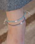Two delicate beaded ankle bracelets on a person's ankle, one in turquoise and white, and one in navy and white, with a small white pearl accent on each. The person is wearing gray denim shorts, and the background is a soft-focus sandy beige.