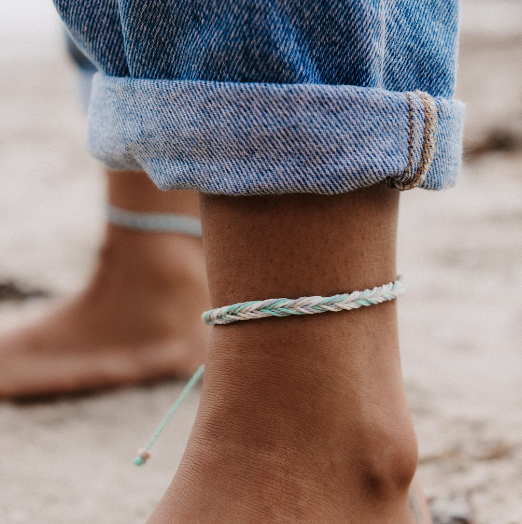 A close-up of a person's bare ankle wearing a thin braided rope anklet in pastel mint green, white, and light blue, with cuffed blue denim jeans visible above it. A second matching anklet is faintly visible on the other ankle in the background.