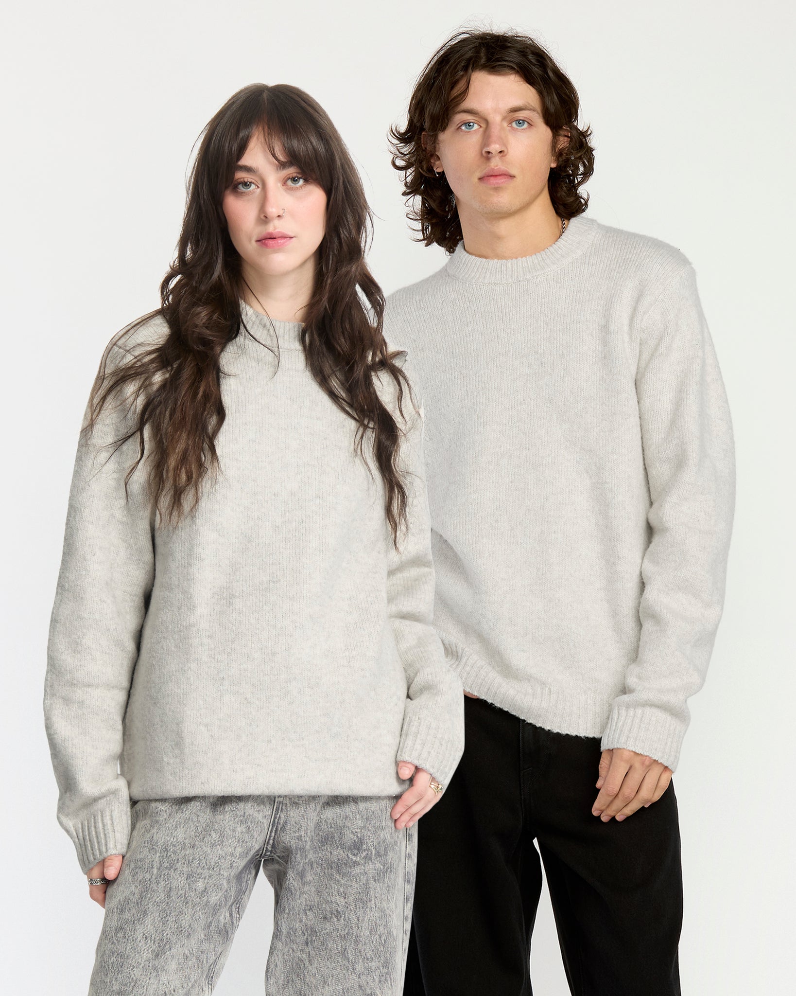 Two people wearing light gray sweaters and dark pants against a white background